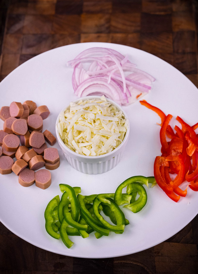 Sliced sausages, shredded cheese, and fresh vegetables for naan pizza toppings.