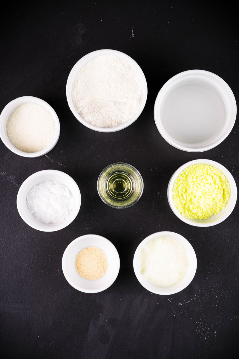 Naan dough ingredients like flour, oil, and yogurt in white bowls on a black background.