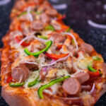 Close-up of cheesy naan pizza topped with sausages, bell peppers, and onions.