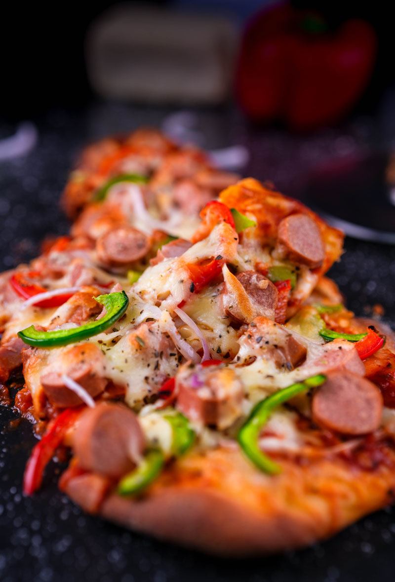 Close-up of naan pizza topped with melted cheese, sausages, bell peppers, and onions.