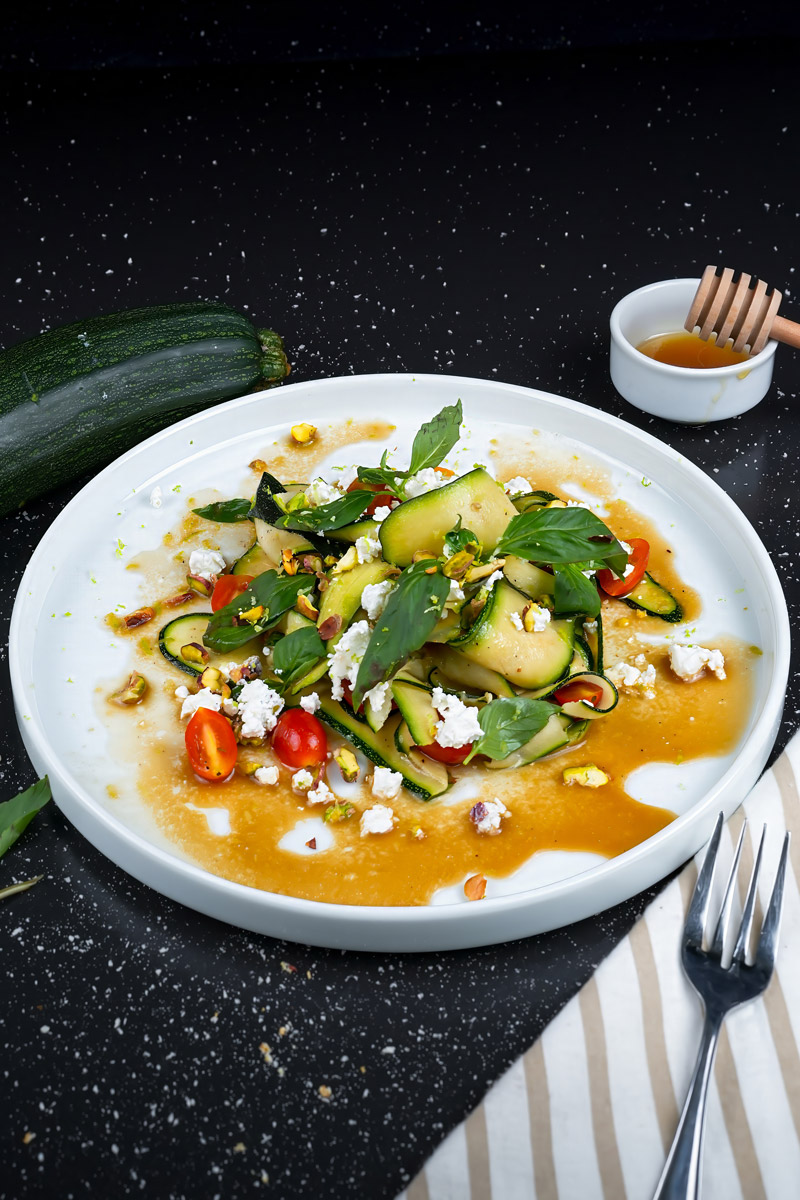 Close-up of raw zucchini salad garnished with fresh herbs, cheese crumbles, and nuts.