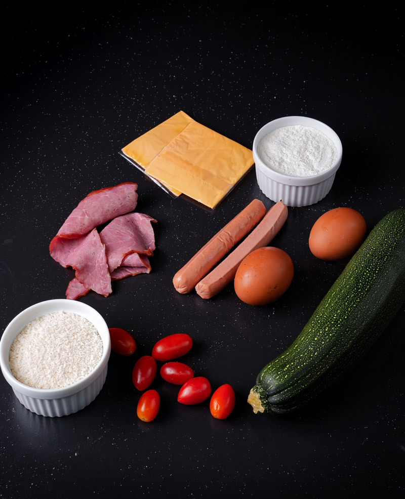 Ingredients for baked zucchini skewers, including zucchini, cheese, eggs, breadcrumbs, and sausages.