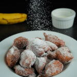 Powdered sugar being sprinkled over banana fritters on a plate