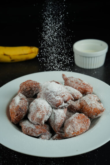 Powdered sugar being sprinkled over banana fritters on a plate