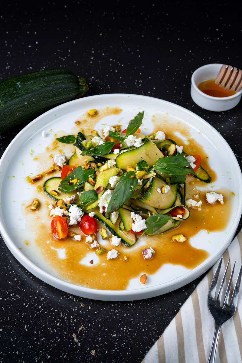 Raw zucchini salad with cherry tomatoes, feta cheese, pistachios, and fresh basil on a white plate.