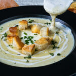 Creamy leek potato soup being served with croutons and fresh herbs.