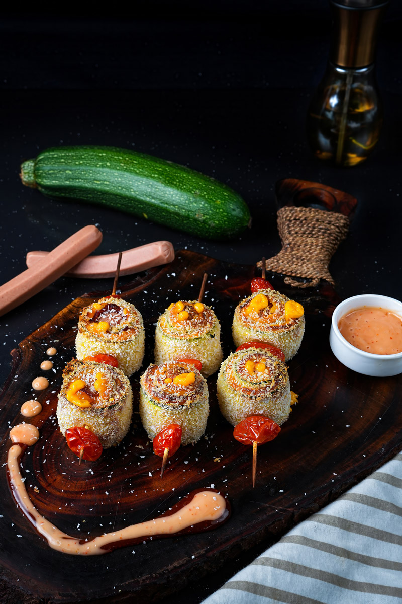 Baked zucchini skewers arranged on a rustic board with sauce and fresh zucchini in the background.