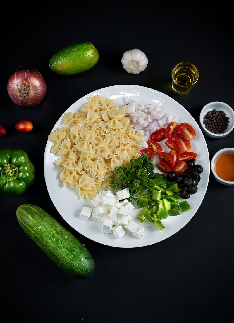 Ingredients for Greek pasta salad laid out on white plate with fresh vegetables