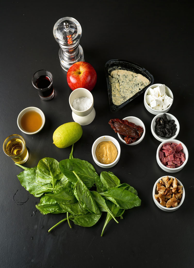 Ingredients for apple pecan salad including spinach, apples, walnuts, and cheese on a black surface