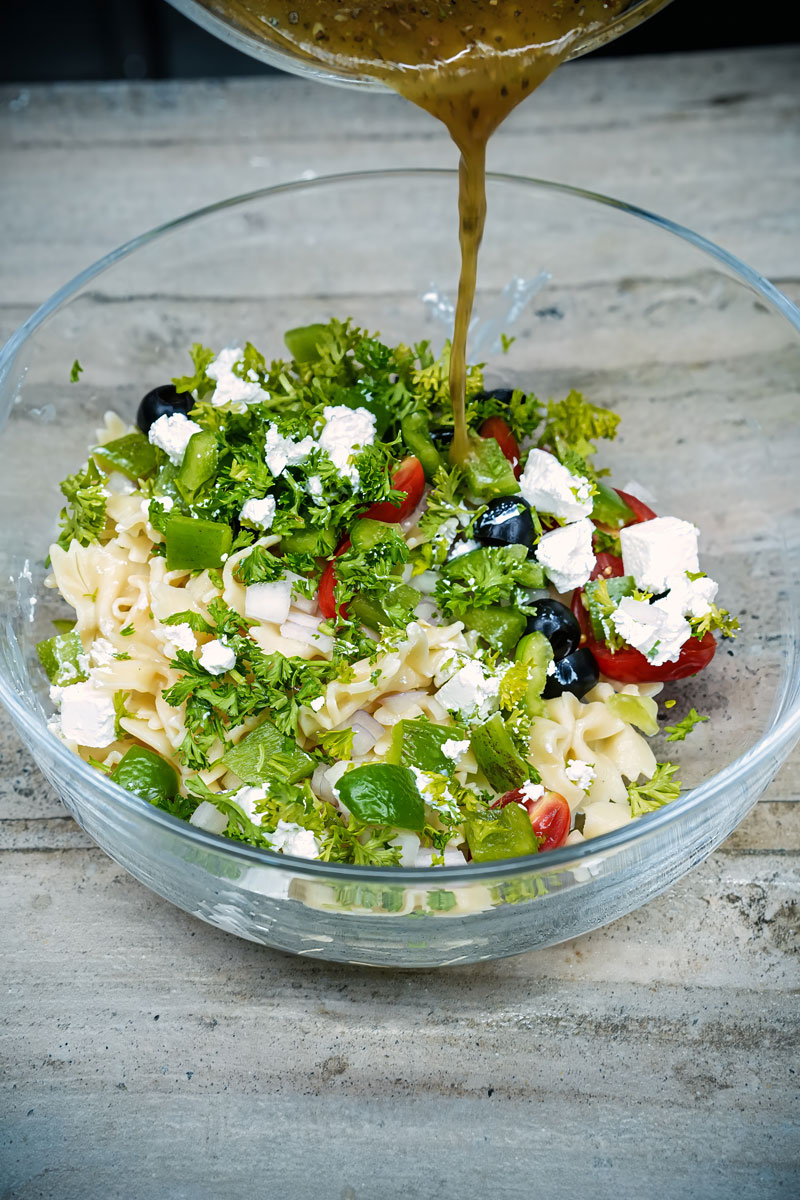 Homemade Greek dressing being poured over farfalle pasta salad with vegetables