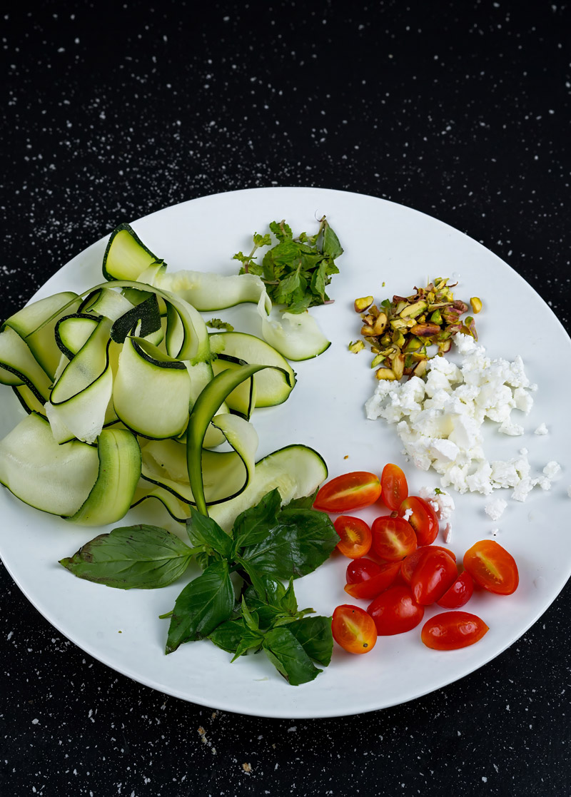 Prepared ingredients for raw zucchini salad arranged on a plate: zucchini ribbons, cherry tomatoes, feta cheese, pistachios, and fresh herbs.