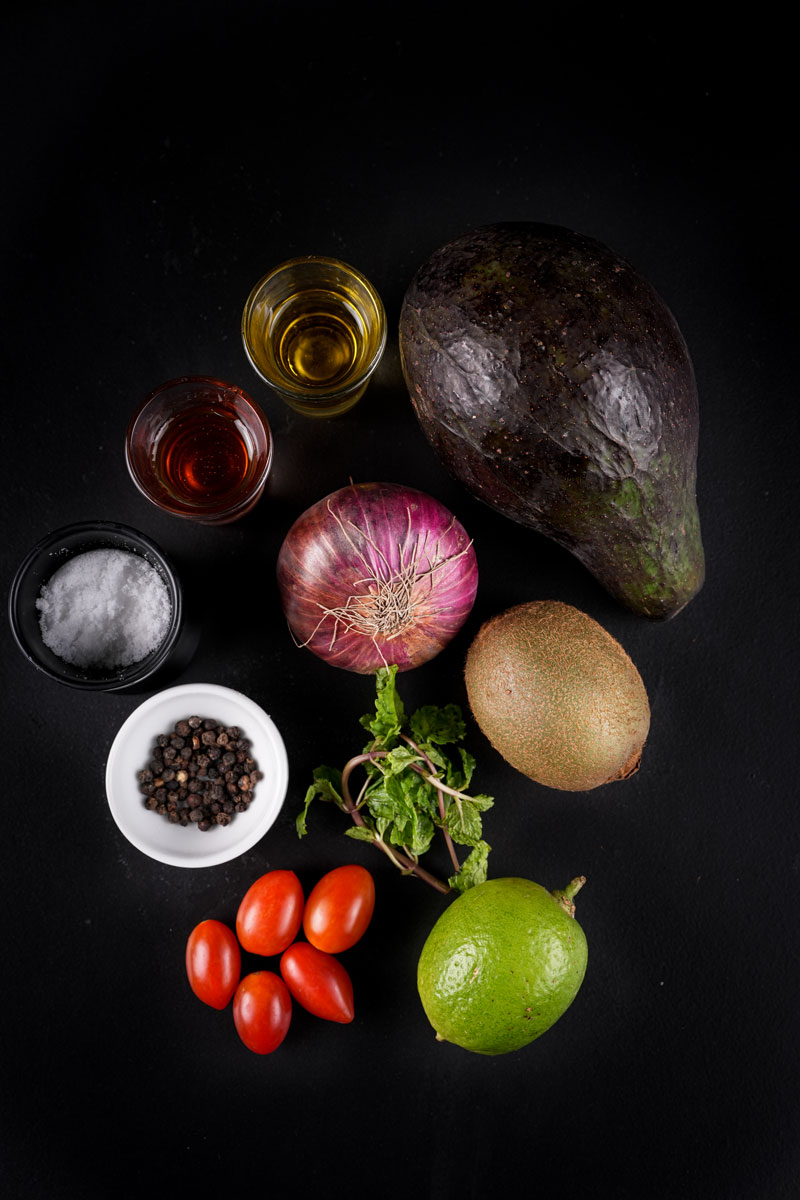 Ingredients for Avocado Kiwi Salad: diced avocado, kiwi, cherry tomatoes, mint leaves, and chopped onions on a white plate.