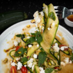 Fork holding a bite of raw zucchini salad with feta cheese and basil.