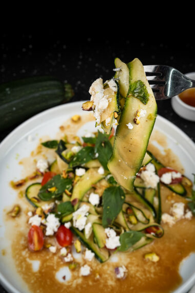 Fork holding a bite of raw zucchini salad with feta cheese and basil.