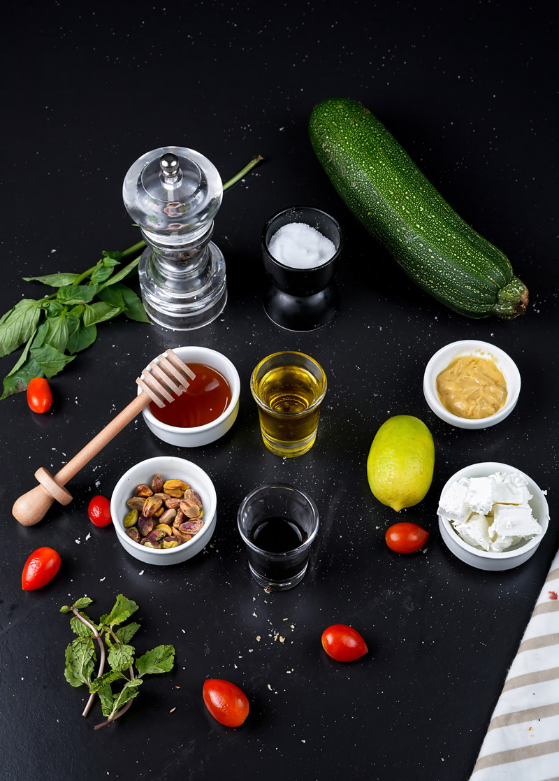 Ingredients for raw zucchini salad including zucchini, cherry tomatoes, feta cheese, pistachios, basil, honey, and lemon.