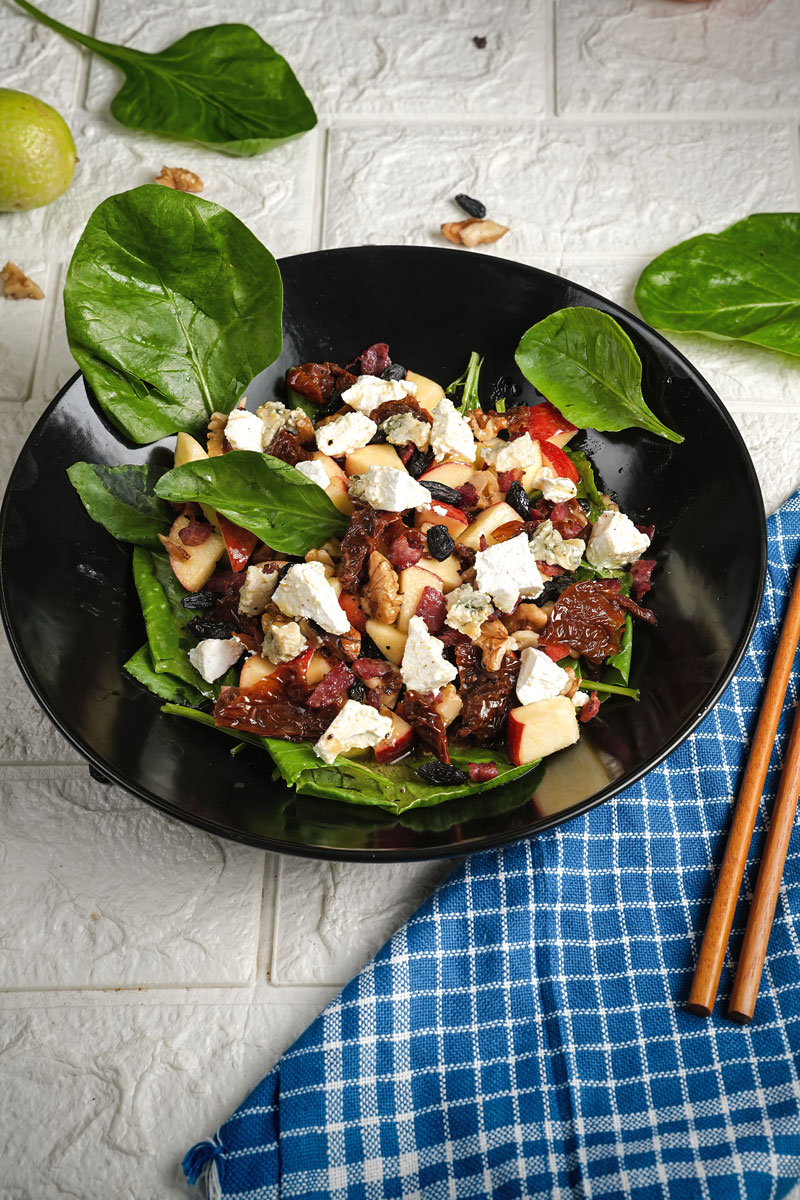 Apple pecan salad with spinach, feta cheese, and sun-dried tomatoes in a black bowl on a white surface