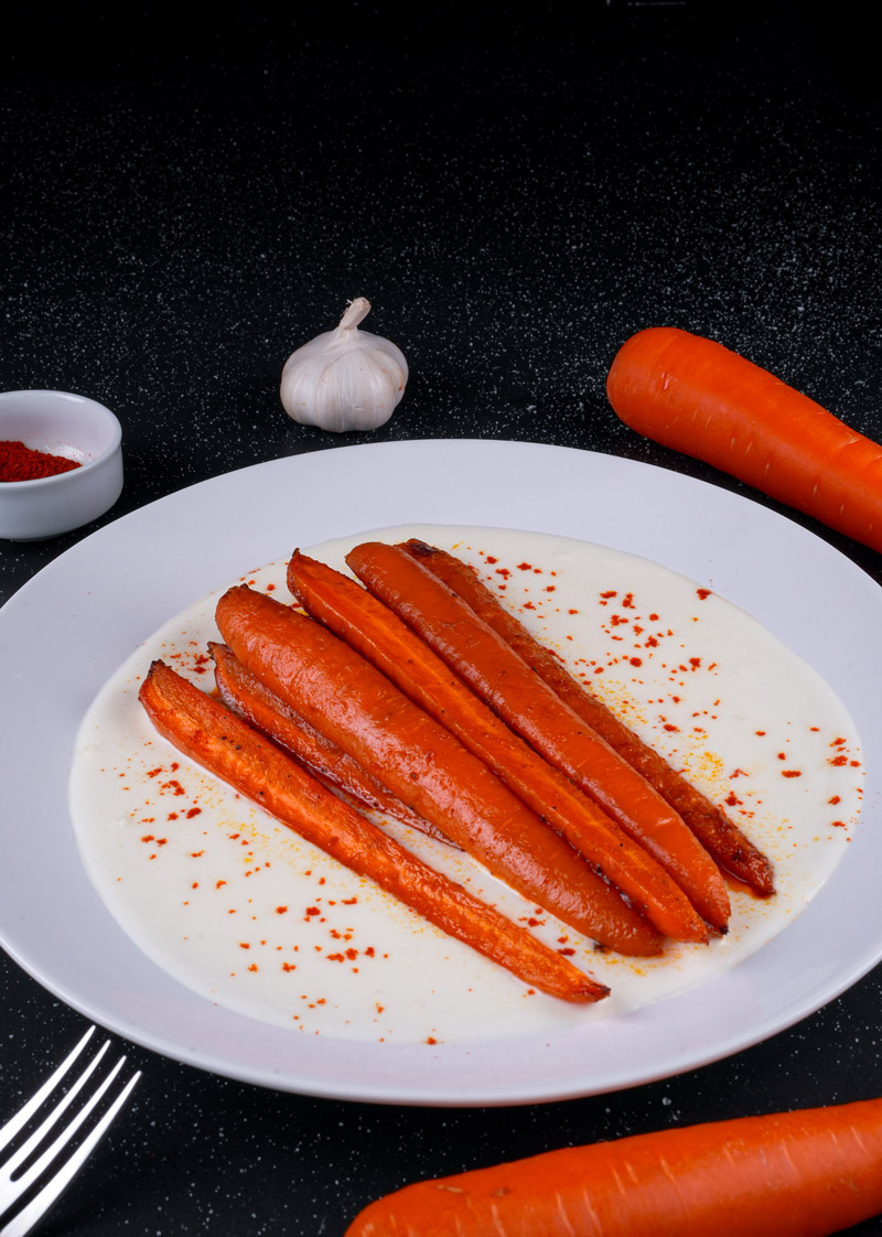 Spicy maple roasted carrots plated with yogurt sauce, surrounded by fresh carrots and garlic.