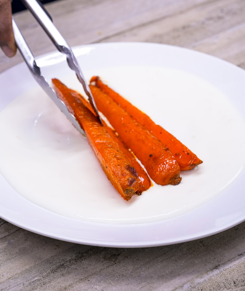 Tongs placing spicy maple roasted carrots on a plate with creamy yogurt sauce.