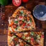 Close-up of Tandoori Paneer Pizza Naan slices on a wooden board with fresh vegetables.