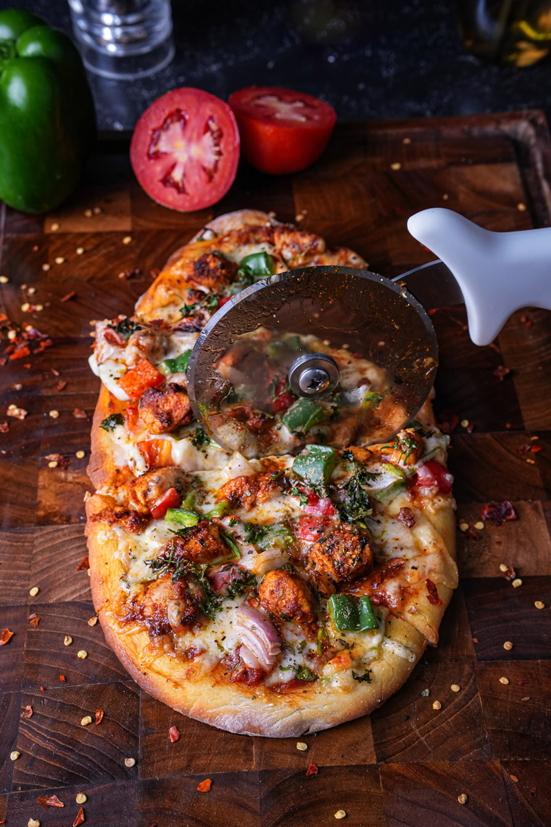 Freshly baked tandoori paneer pizza naan being sliced with a pizza cutter on a wooden board