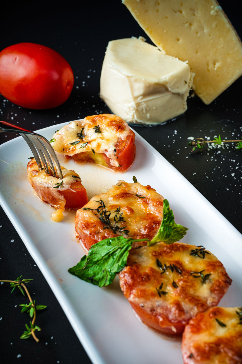 Close-up of baked tomatoes with cheese, garnished with herbs and served with a fork.