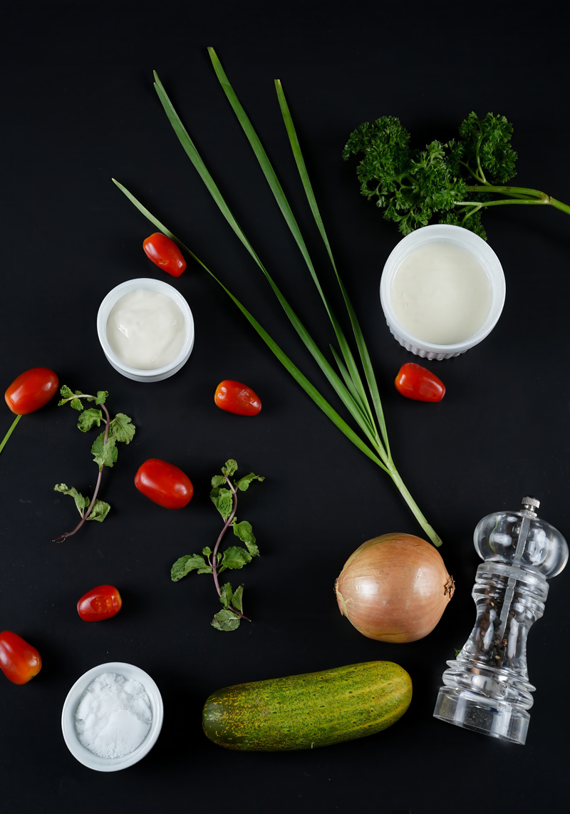 Ingredients for creamy cucumber tomato salad laid on a dark background.