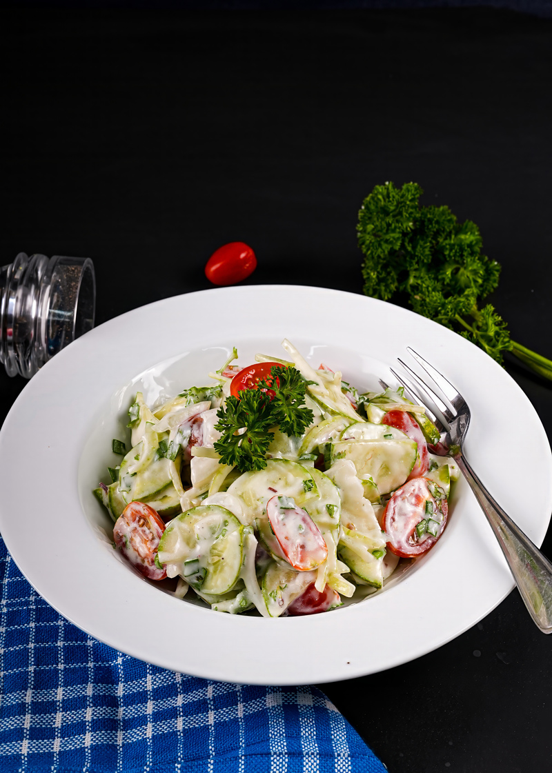 Top view of cucumber tomato salad in a white bowl on a checkered blue napkin.