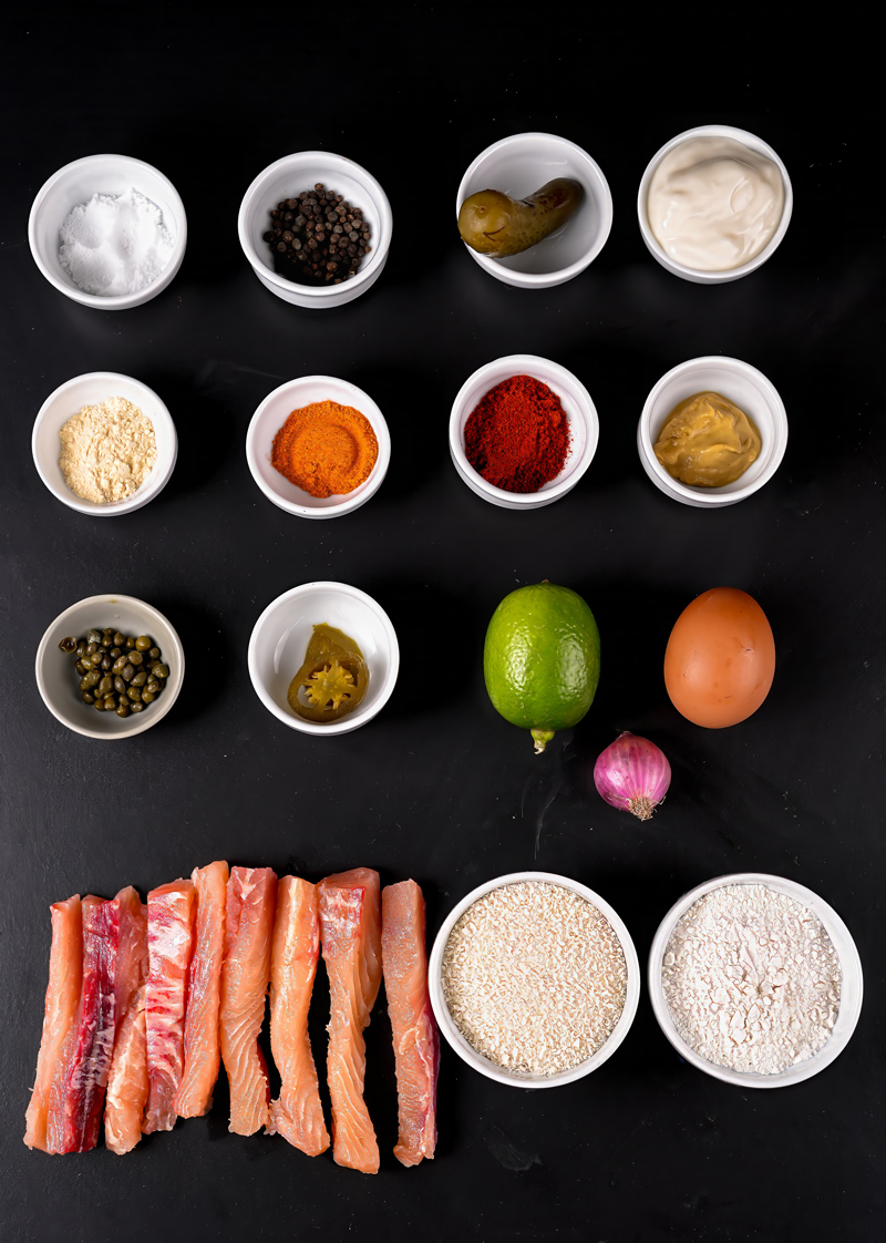 Ingredients for homemade fish sticks arranged in small bowls on a black surface.