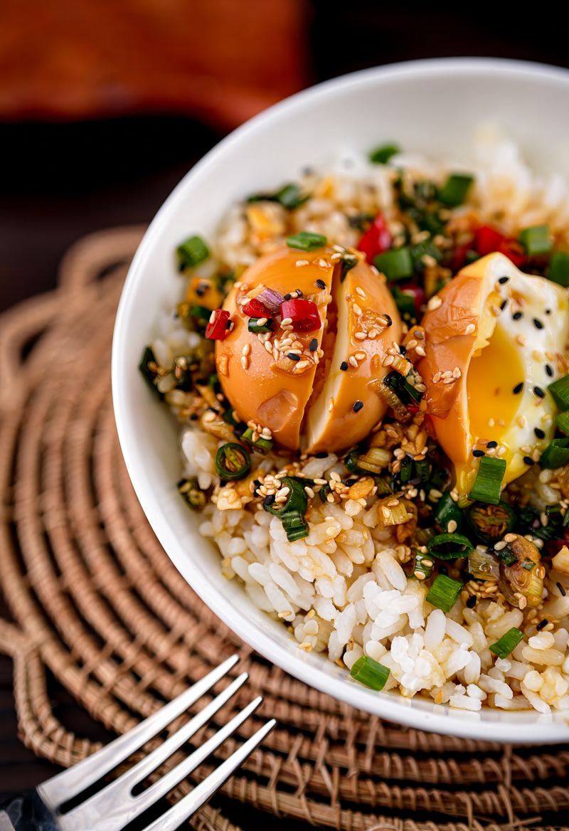 Overhead view of a bowl of rice topped with sliced Mayak Eggs, sesame seeds, and chili garnish.