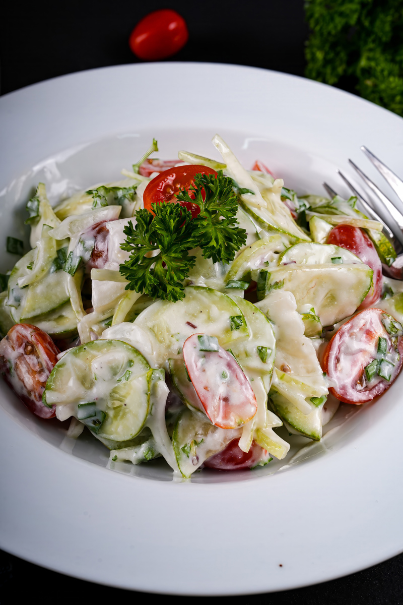 Fresh creamy cucumber tomato salad garnished with parsley on a dark backdrop.