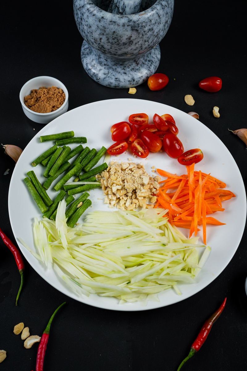 Chopped ingredients for Green Papaya Salad arranged on a white plate with a mortar and pestle in the background.