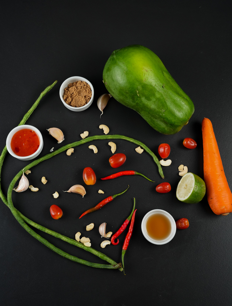 Ingredients for Green Papaya Salad including green papaya, carrots, chilies, lime, and garlic on a black background.