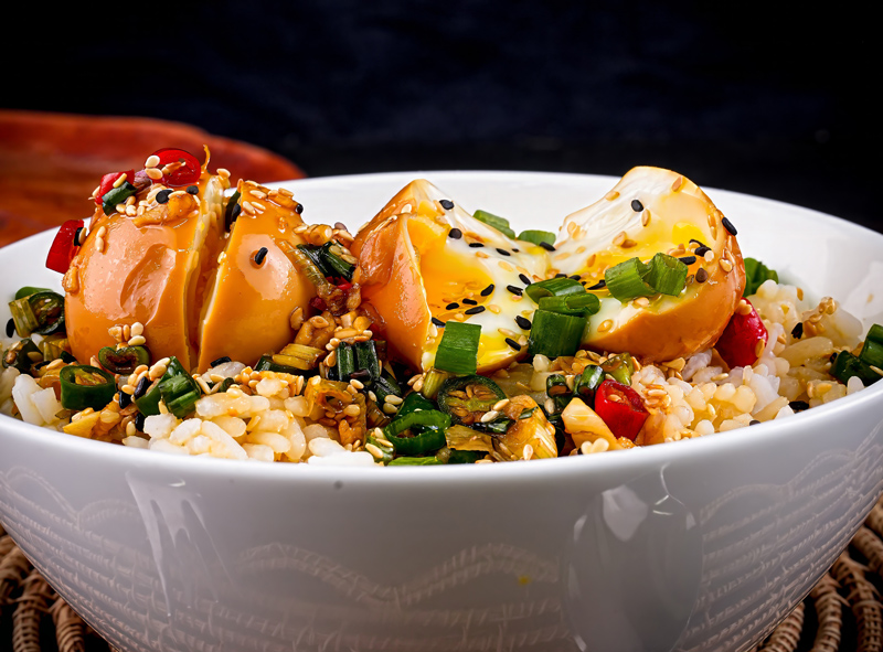 Top-down view of Mayak Eggs garnished with sesame seeds, chilies, and green onions on rice.