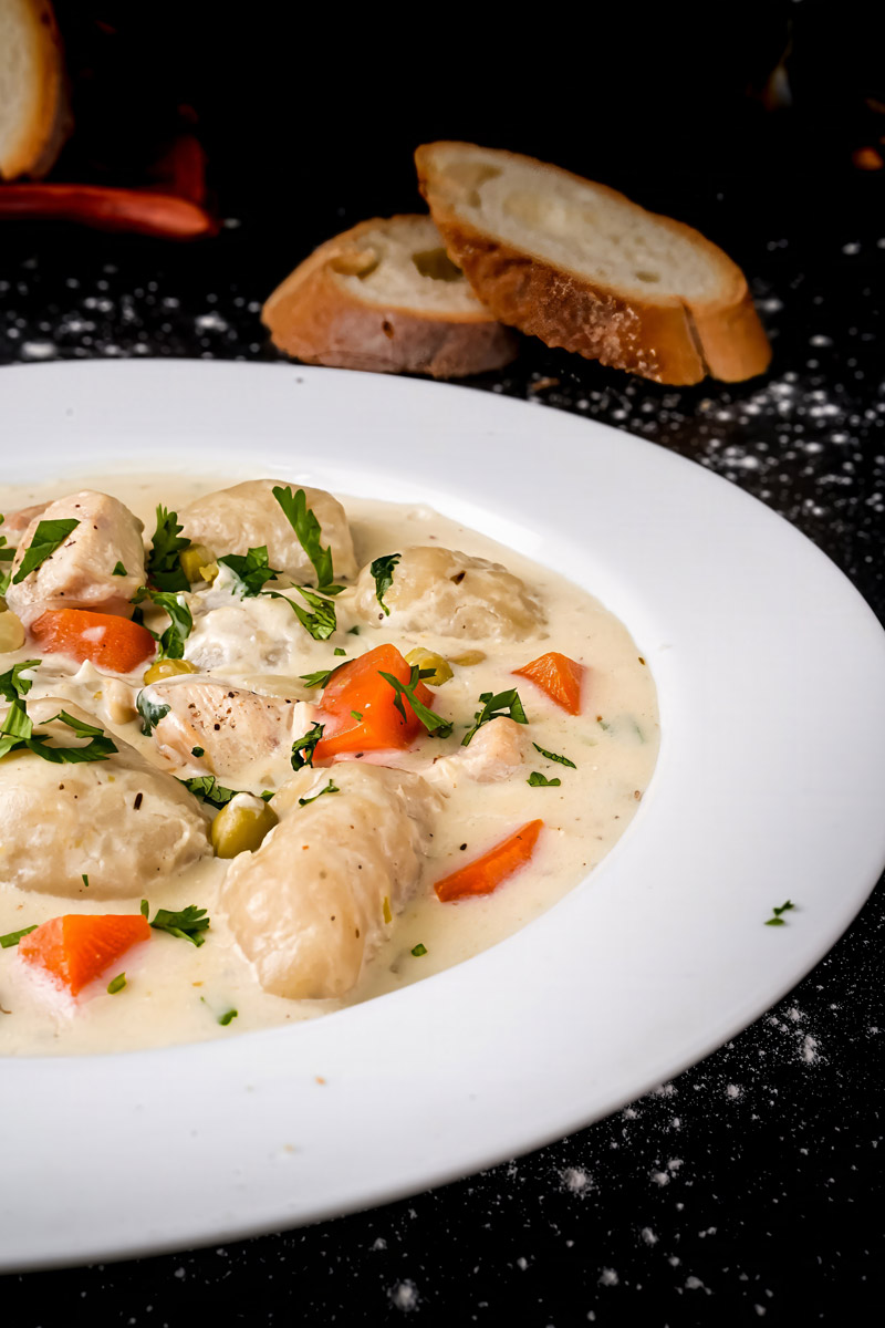 Close-up of creamy chicken and dumplings with fresh herbs and vegetables in a white plate.