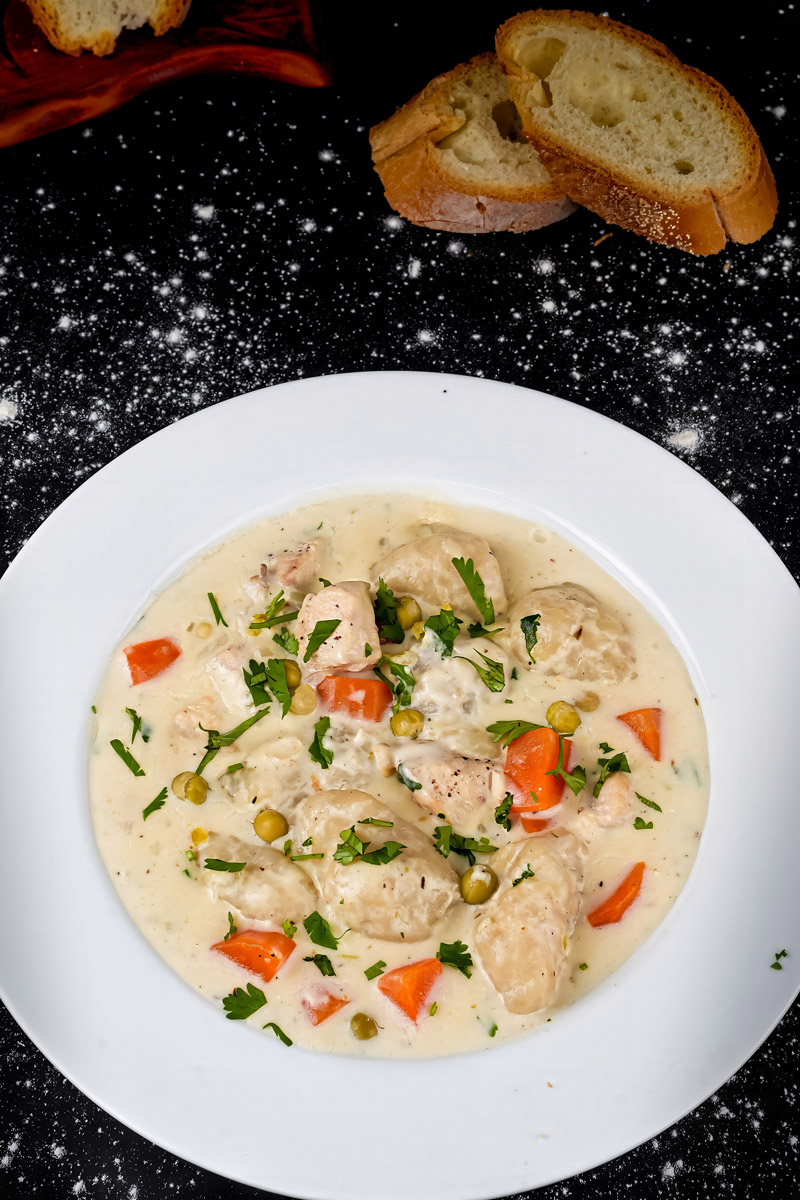 Top-down view of creamy chicken stew with dumplings, carrots, peas, and fresh parsley in a white bowl.