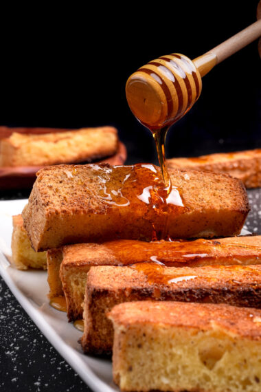 Honey drizzling over golden baked French toast sticks on white serving plate