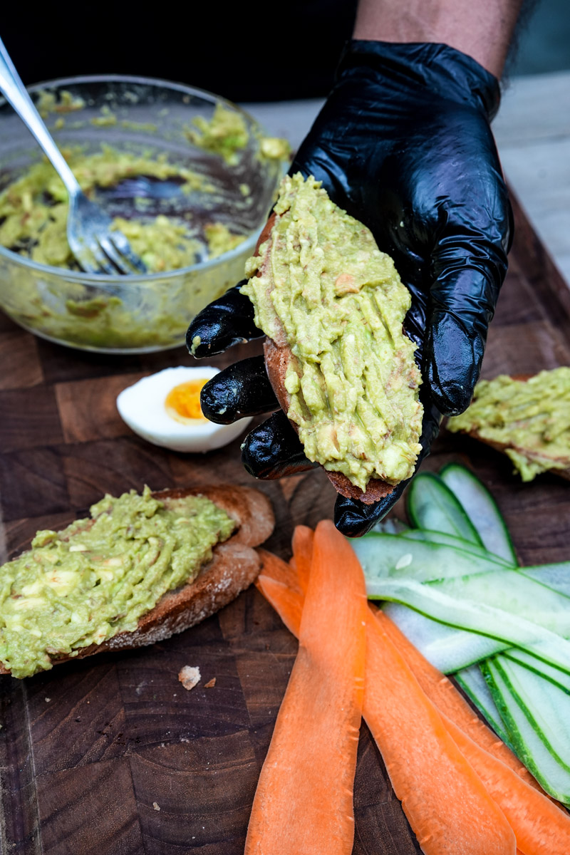 Spreading avocado on toast with gloved hands and prepped ingredients