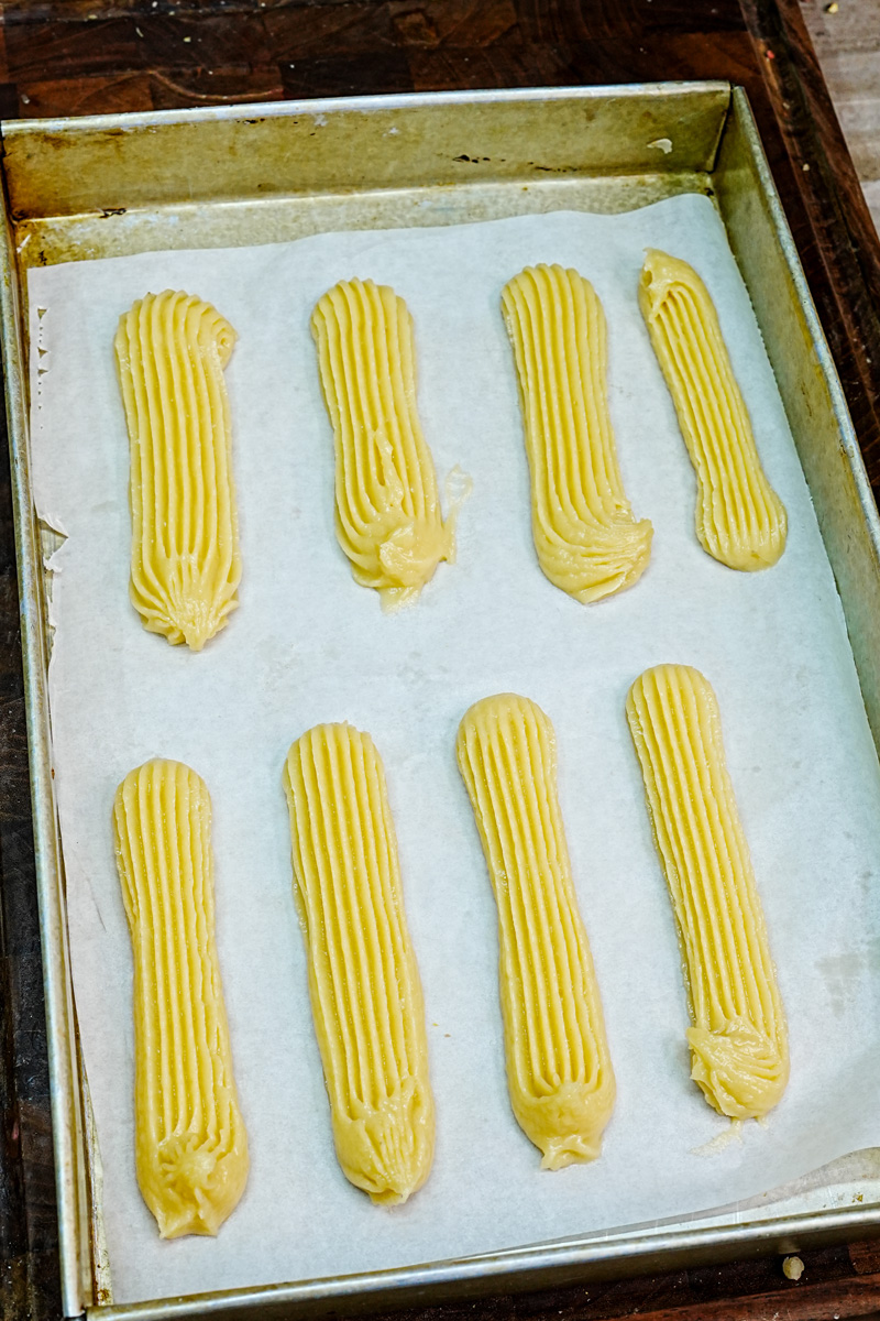 Raw eclair shells piped on baking sheet before baking in oven