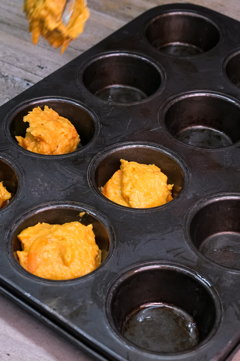 Spooning mango muffin batter into greased muffin tin