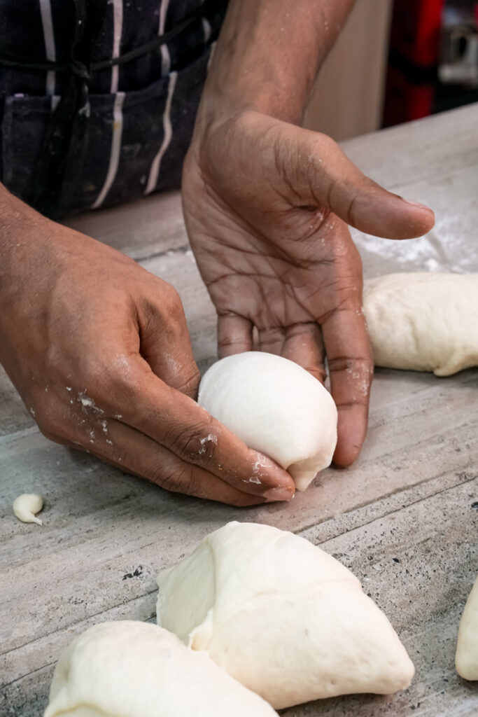 Hands shaping a portion of dough into a smooth ball for a bread bowl.