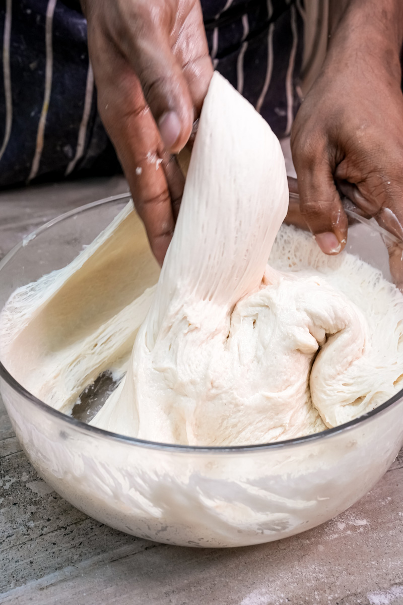 Stretching the wet, sticky dough for English Muffin Bread in a glass bowl.