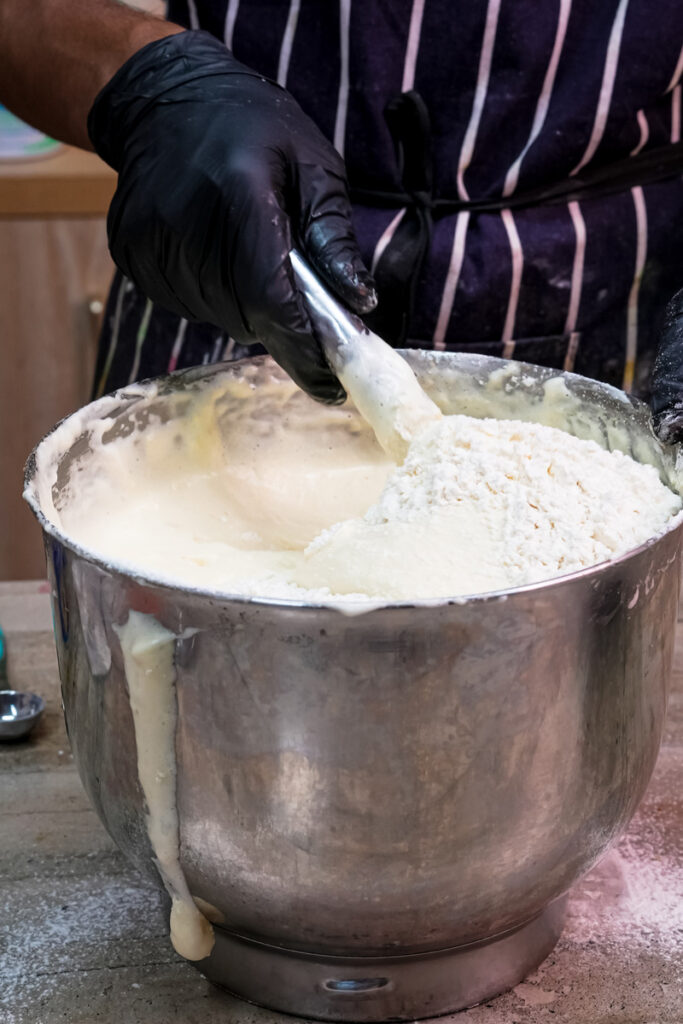A baker adding flour to a bowl of cake batter while wearing black gloves
