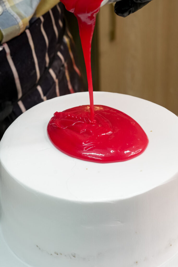 A baker pouring red glaze onto the top of a white frosted cake