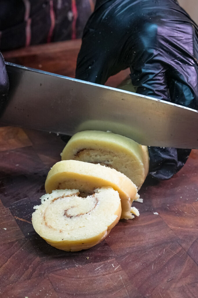A knife slicing a chilled cinnamon roll cookie dough log into individual rounds