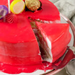A slice of red drip cake being removed from the main cake on a cake stand