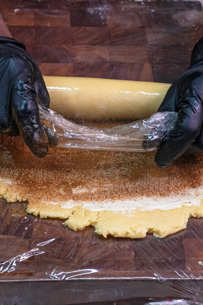 Hands rolling the cinnamon and sugar-coated dough into a log using plastic wrap