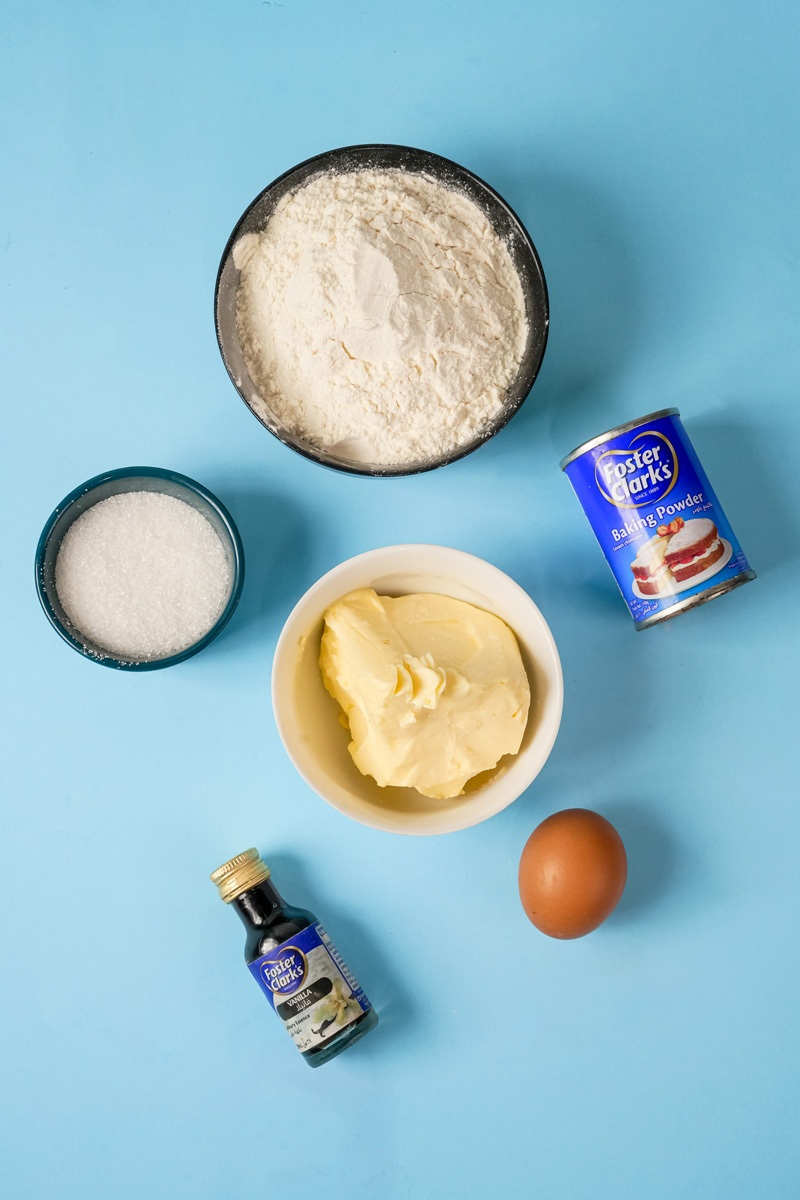 Ingredients for Cinnamon Roll Cookies: flour, baking powder, sugar, butter, vanilla extract, and an egg on a blue background