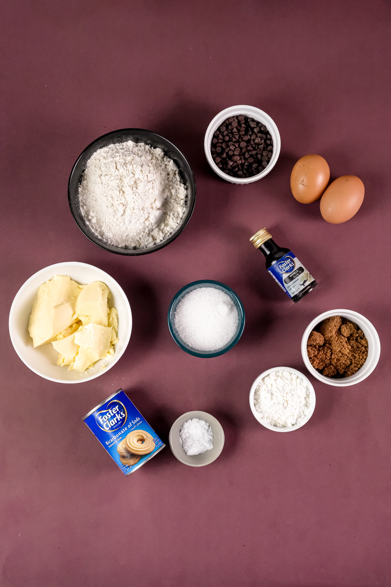 Ingredients for Crumbl Cookies including flour, sugar, butter, eggs, and chocolate chips on a brown surface.