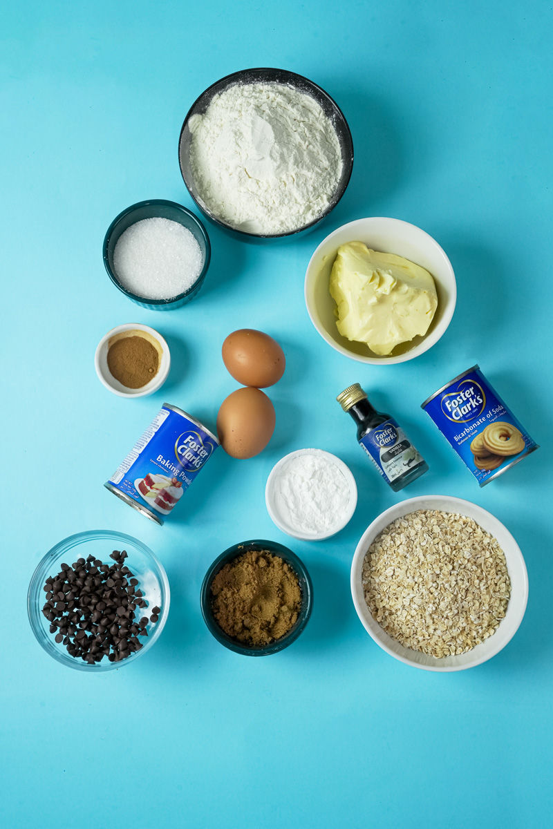 Ingredients for baking oatmeal cookies laid out on a blue background, including oats, flour, sugar, eggs, and chocolate chips