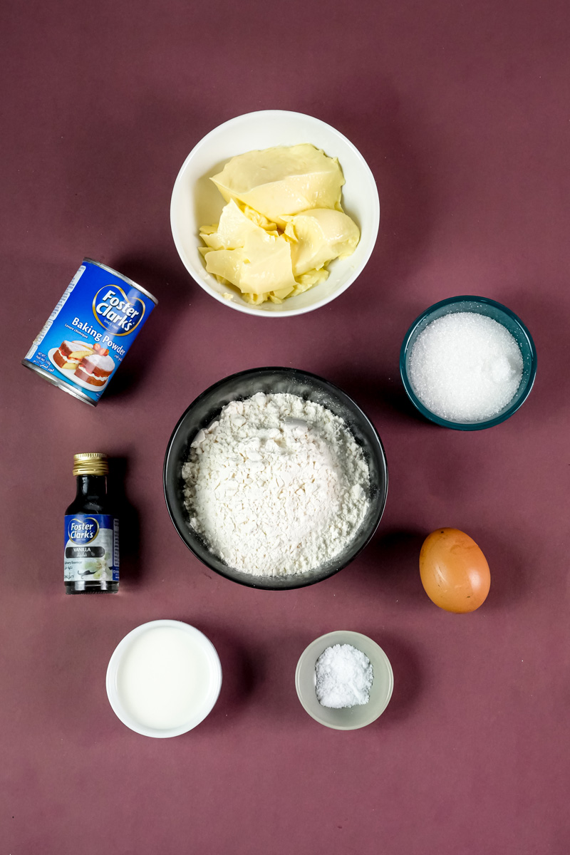 Ingredients for churro cookies on a maroon background, including flour, butter, sugar, an egg, baking powder, vanilla extract, and milk.